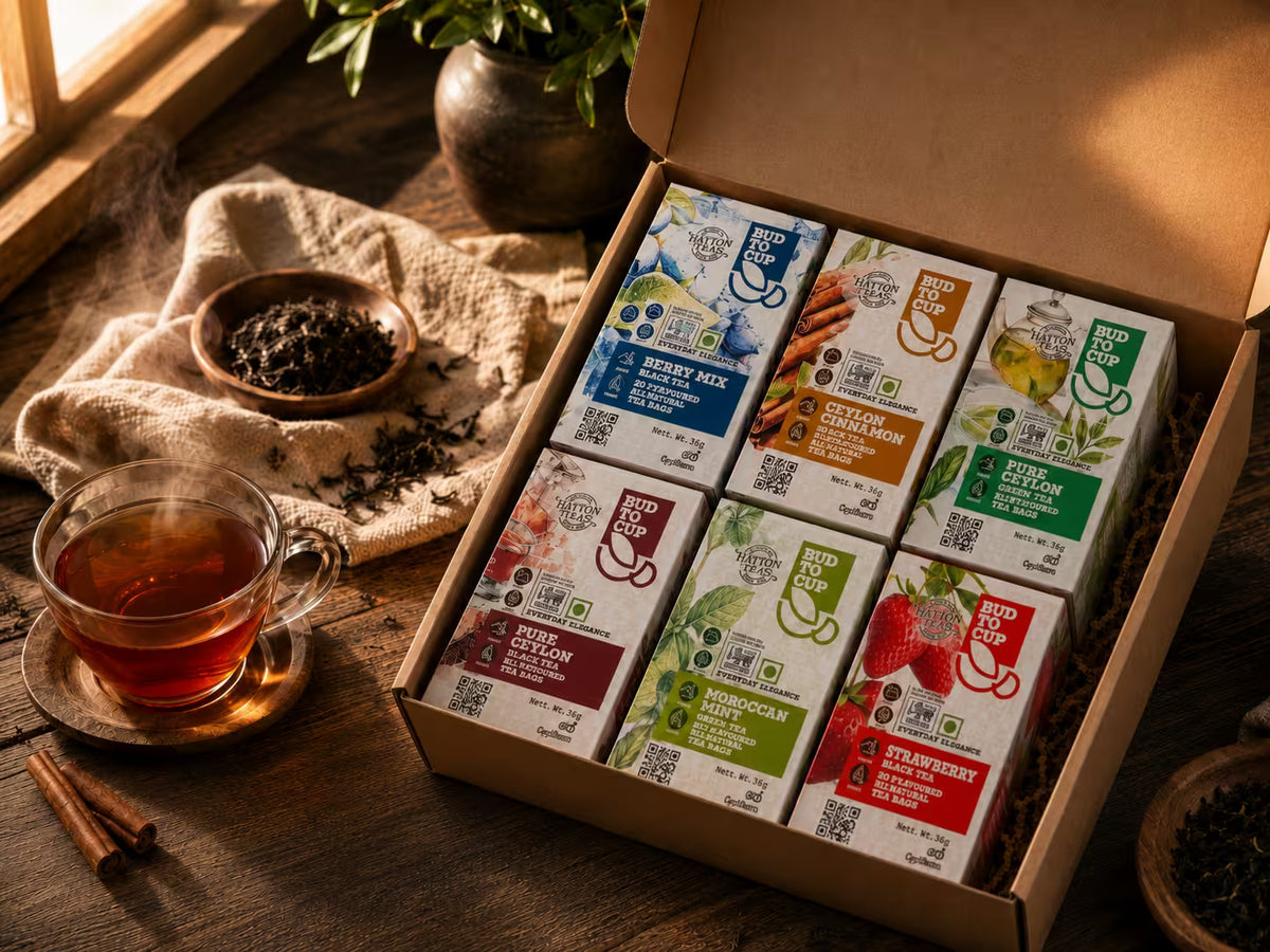 An open gift box filled with six Bud To Cup Ceylon tea packs in assorted flavors, styled on a rustic wooden table with a glass of tea, loose tea leaves, and a plant in the background.